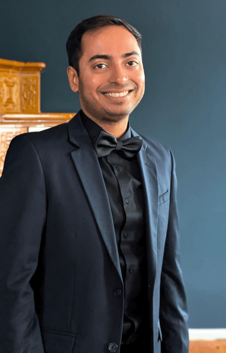 Smiling man in a formal black suit and bow tie against a dark blue background.